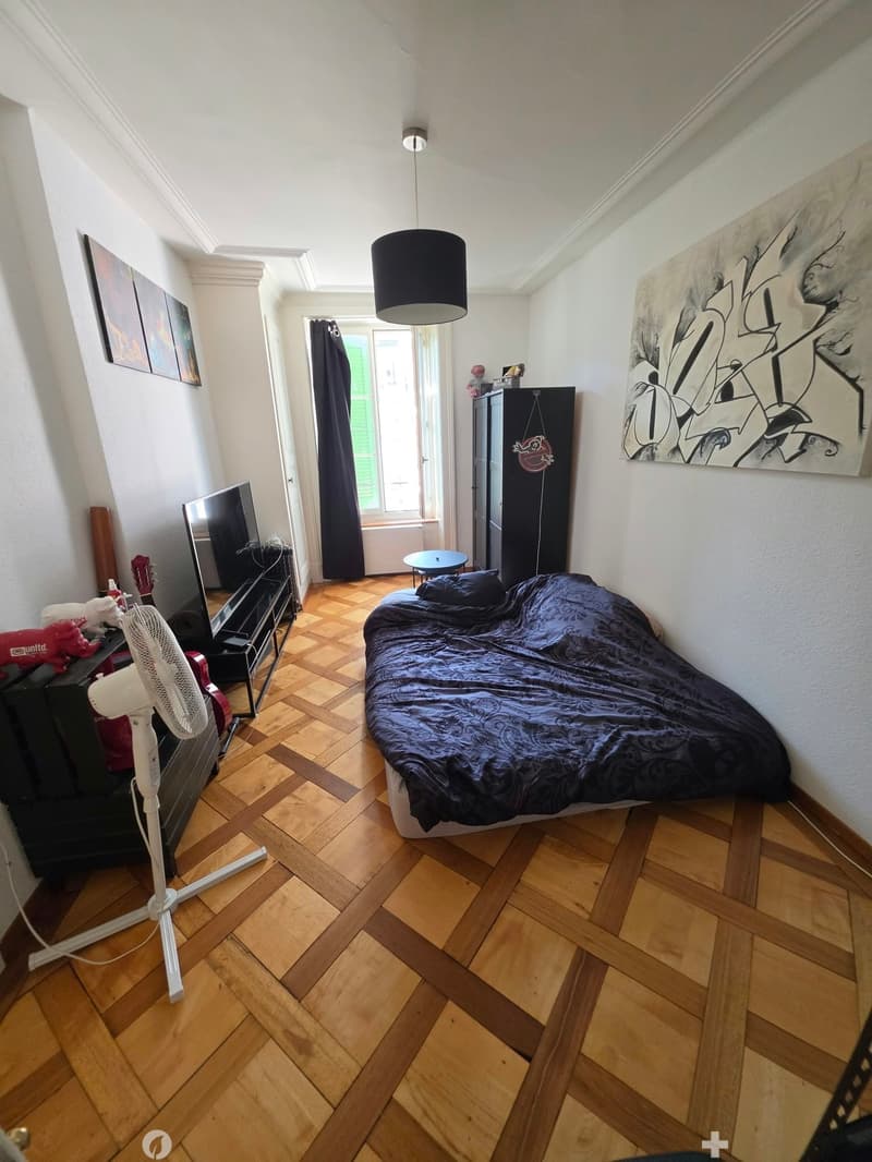 A furnished bedroom with a wooden floor, a bed with dark bedding, a fan, a guitar, a TV stand, a small table, and a large window with dark curtains.