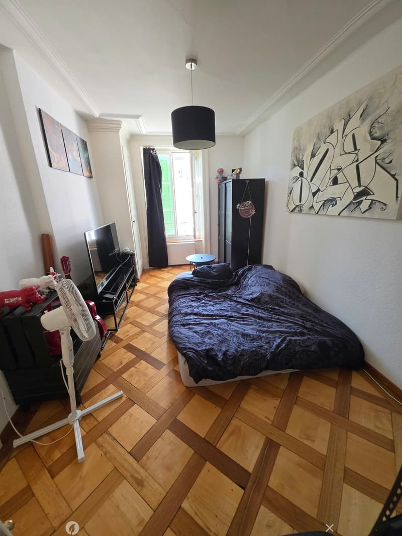 A furnished bedroom with a wooden floor, a bed with dark bedding, a fan, a guitar, a TV stand, a small table, and a large window with dark curtains.