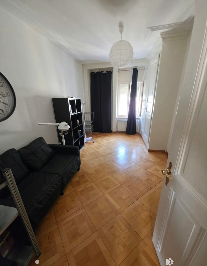 A furnished room with wooden flooring, a black sofa, a shelf, a clock, and a hanging lamp.