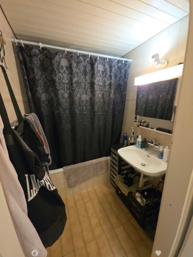 Bathroom with a sink, mirror, shower curtain, and a backpack hanging on the wall. The floor is tiled, and there is a towel on the edge of the bathtub.