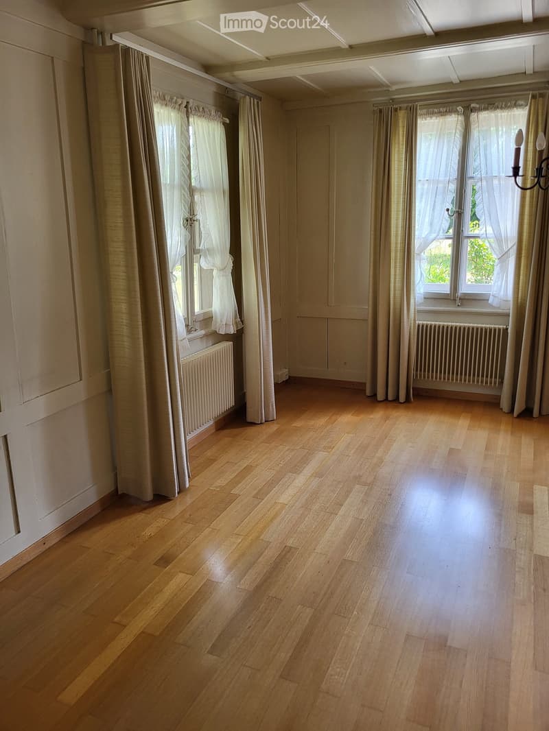 Empty room with wooden flooring, large windows with curtains, radiators, and a chandelier on the wall.