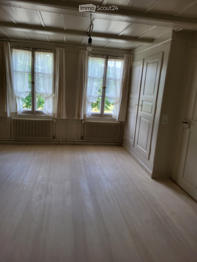 Empty room with wooden flooring, large windows with white curtains, radiators, and white walls.