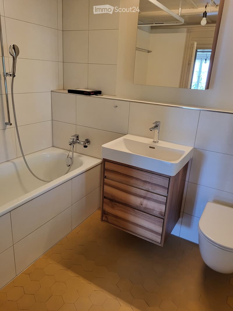 Modern bathroom with a white bathtub, a white sink with wooden cabinet, a toilet, and a mirror above the sink. The walls are tiled in light beige, and the floor is covered with hexagonal tiles.