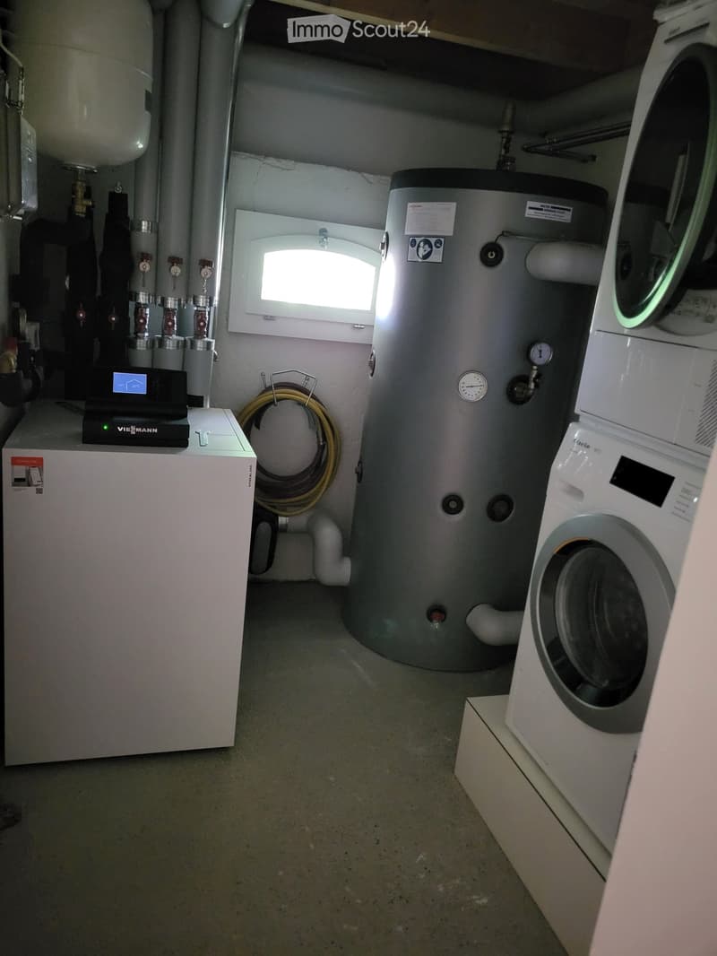 Utility room with a washing machine, water heater, and control panel, featuring a small window and various pipes.