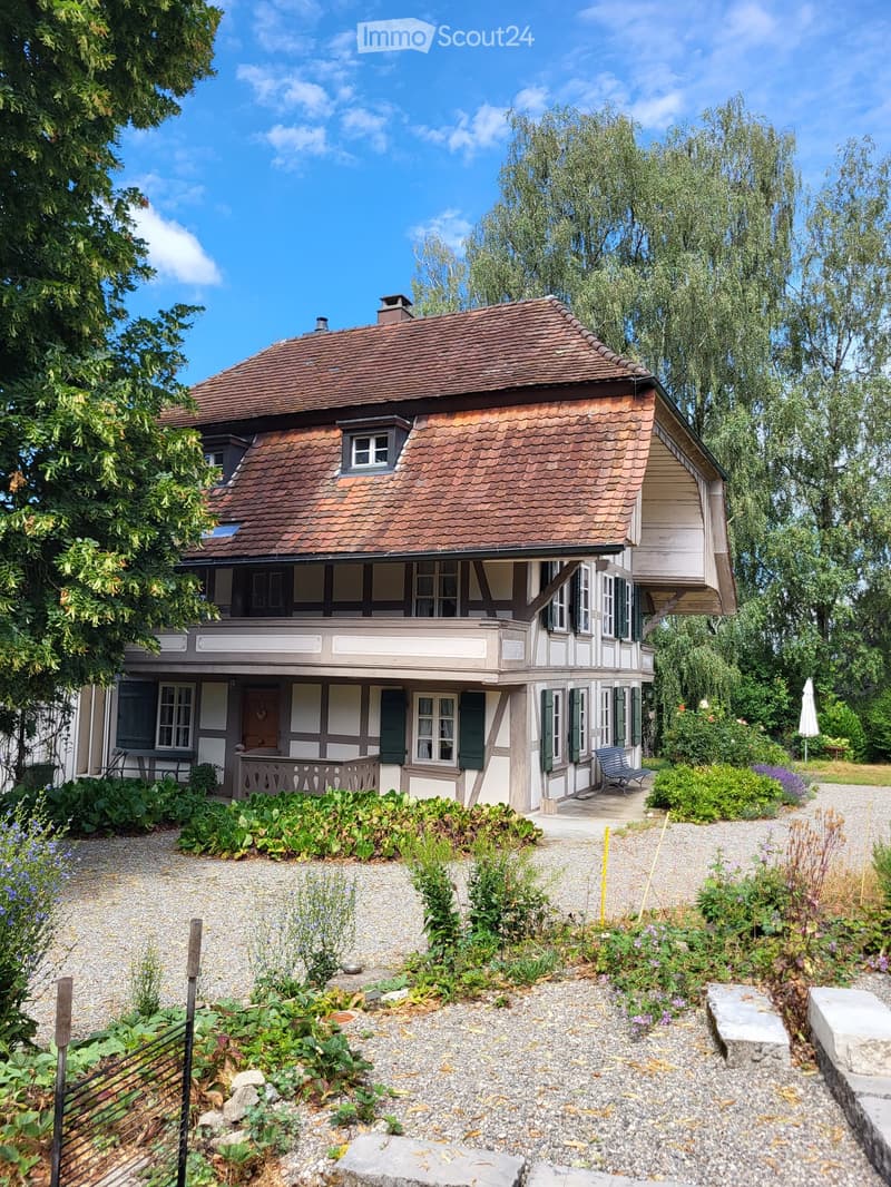 Two-story traditional house with a brown tiled roof, white and grey exterior, multiple windows with shutters, and a well-maintained garden with gravel pathways and various plants.