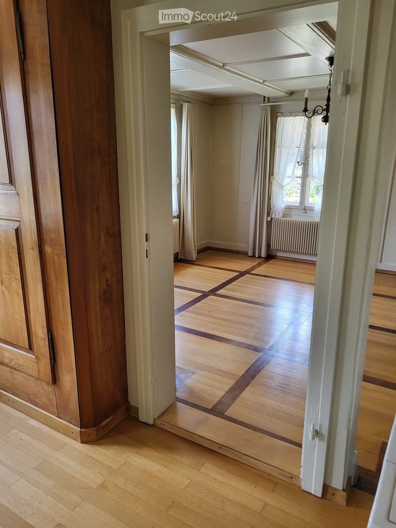 Empty room with wooden flooring, white walls, and large windows with curtains. The room is connected to a hallway with wooden doors and a chandelier visible in the adjacent space.