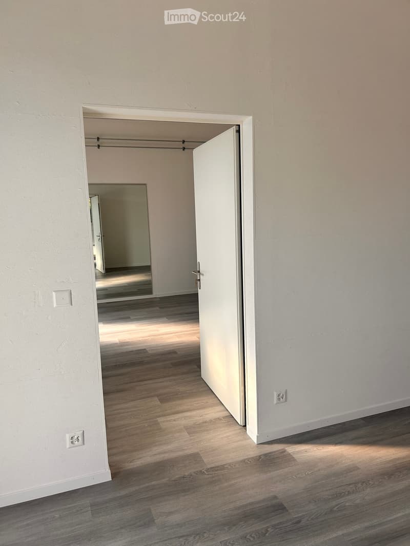 Empty hallway with light wooden flooring and white walls, featuring an open door leading to another room with a visible closet inside.