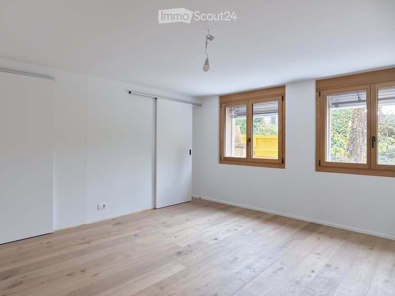 Empty room with light wooden flooring, white walls, and large windows with wooden frames. A sliding door is visible on the left side.