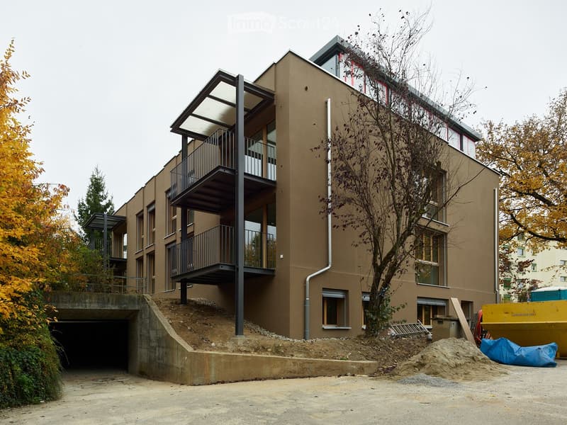 Modern multi-story building with beige facade, large windows, and balconies. Features a raised ground floor with a garage entrance and construction materials nearby.