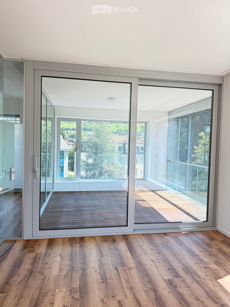 Empty room with wooden flooring, large sliding glass doors leading to a balcony with wooden decking and a scenic view of greenery and houses.