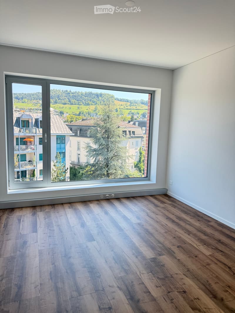 Empty room with wooden flooring and large window offering scenic view of greenery and buildings.