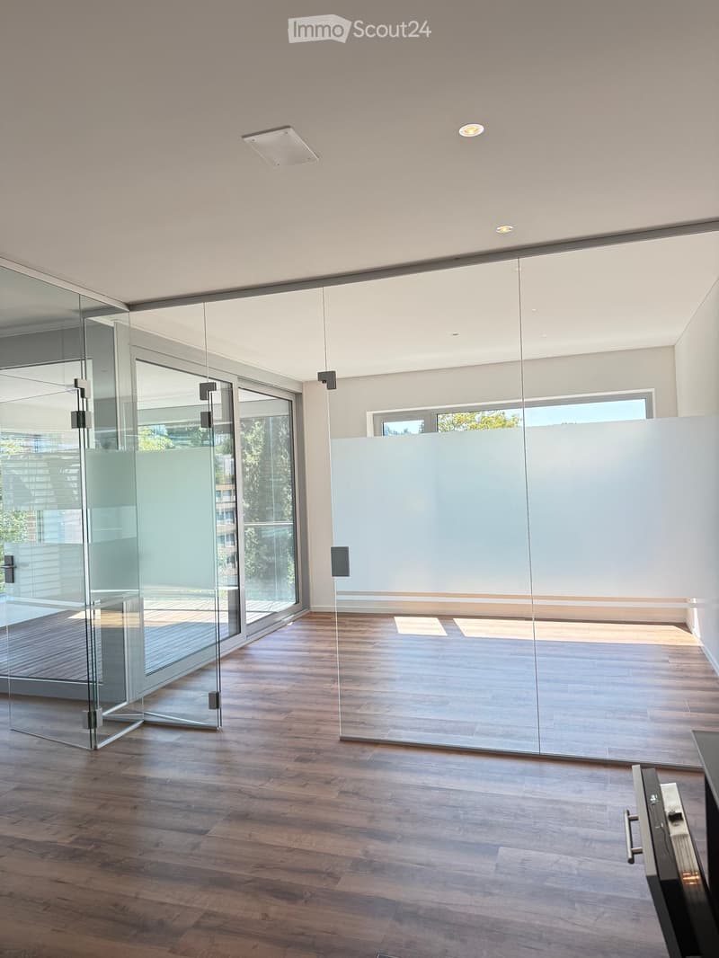 Modern, minimalist room with wooden flooring, large glass walls, and recessed lighting. The space is completely empty with no furniture or furnishings.
