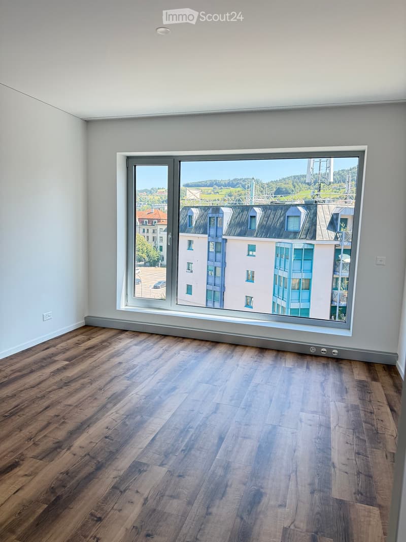Empty room with wooden flooring and large window offering a view of the cityscape and surrounding buildings.