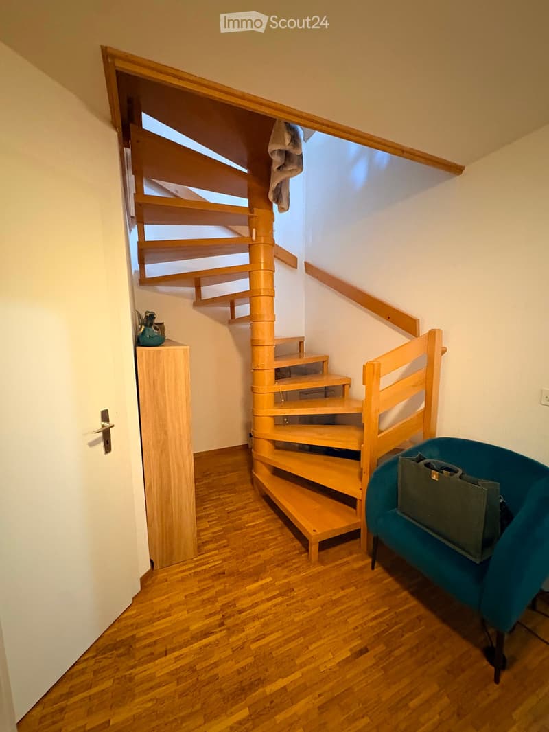 A small, cozy room with wooden flooring, a spiral staircase leading upwards, a small wooden cabinet, and a teal-colored sofa with a handbag placed on it.