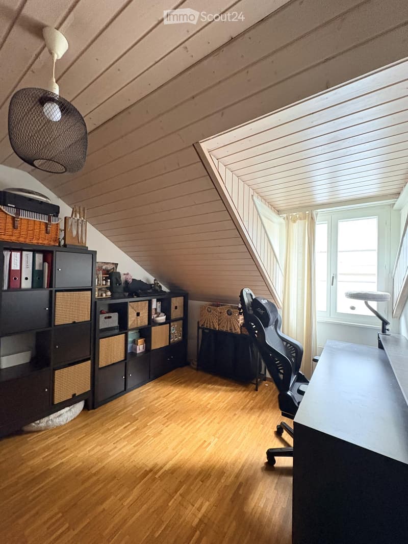 Attic room with wooden flooring, slanted wooden ceiling, large window with white curtain, black storage unit with wicker baskets, desk, office chair, and a hanging fan.
