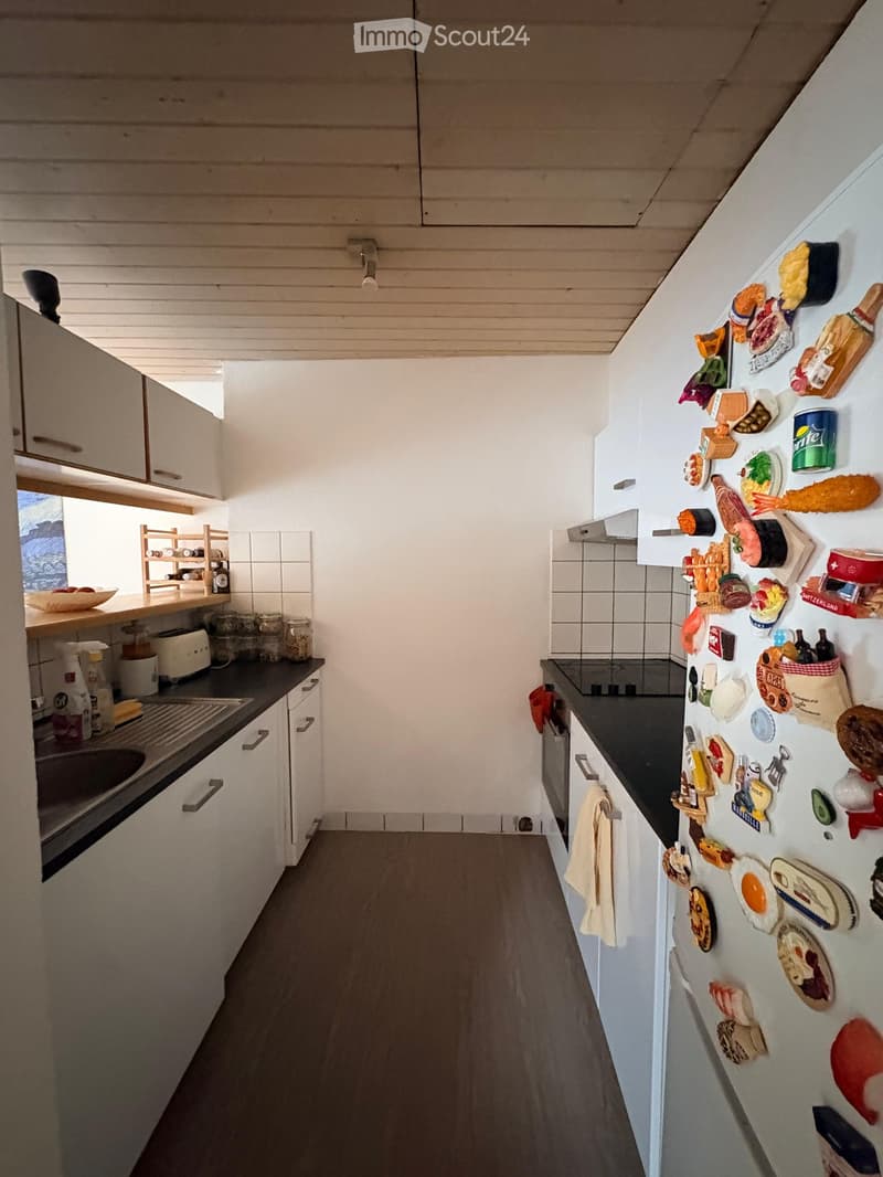 Modern kitchen with white cabinets, dark countertops, and a refrigerator adorned with various magnets and decorations. Includes a sink, toaster, and a small wooden shelf with jars and bottles.