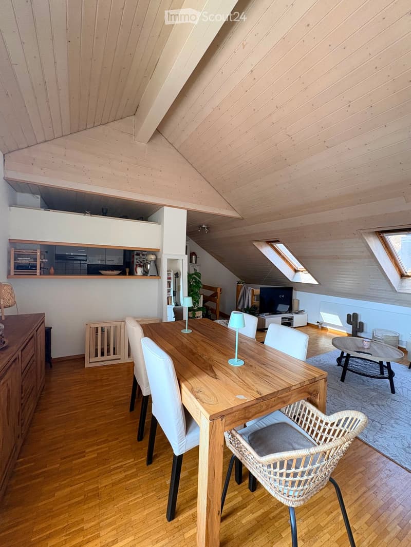 Bright, open-plan living and dining area with wooden floors, slanted wooden ceiling, large windows, and skylights. Features a wooden dining table with white chairs, a wicker chair, a small coffee table, a TV, and a kitchen area with cabinets and appliance