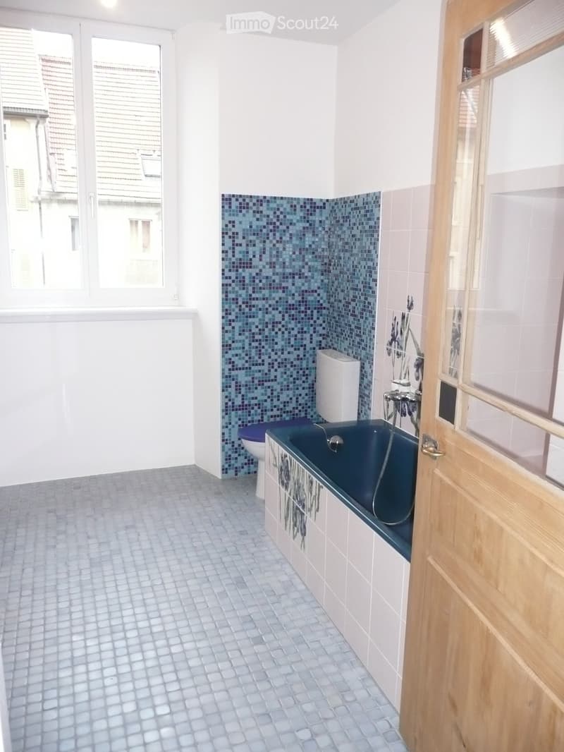 Bathroom with a dark blue bathtub, white toilet, mosaic tile accent wall, and light grey tiled floor.