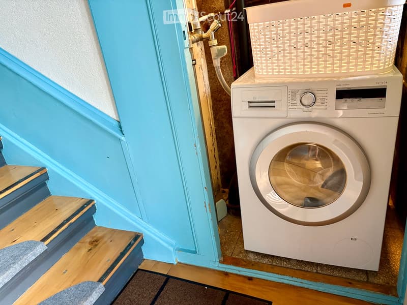 A compact bathroom with a white Bosch washing machine and a woven basket on top, featuring a blue-painted wooden door frame and tiled floor.