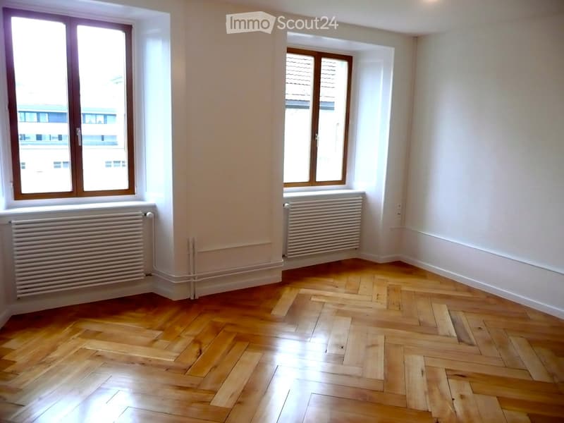 Empty room with wooden herringbone flooring, white walls, and three windows with wooden frames. Radiators are present under two of the windows.