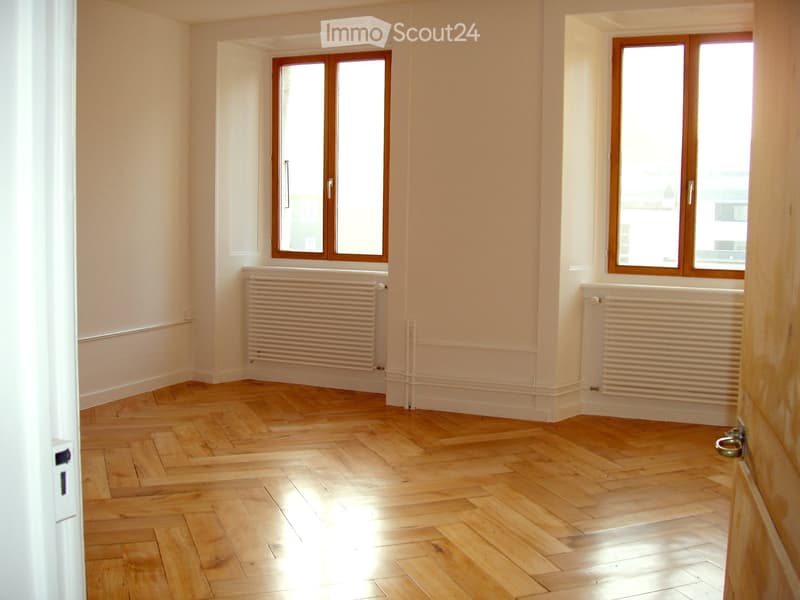 Empty room with wooden herringbone flooring, white walls, and two windows with wooden frames. Radiators are present under each window.