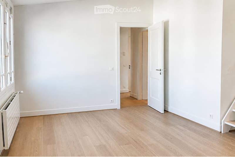 Empty room with light wooden flooring, white walls, and a window with white frames. A radiator is visible on the left side, and an open door leads to another room.
