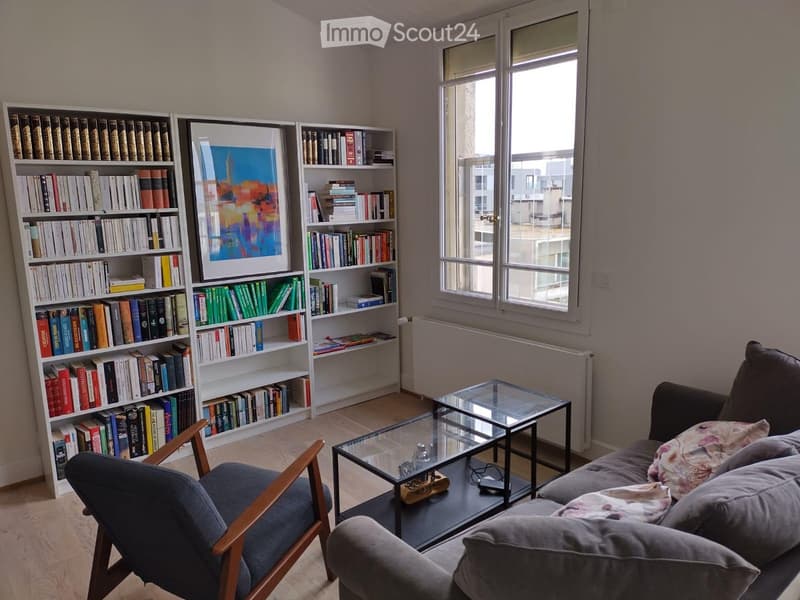 Living room with a grey sofa, wooden armchair, glass coffee table, bookshelf filled with books, and a framed picture on the wall.