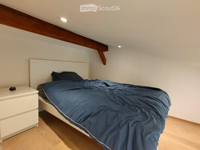 Cozy bedroom with a wooden bed frame, white headboard, blue duvet, and a white nightstand with a small object on top.