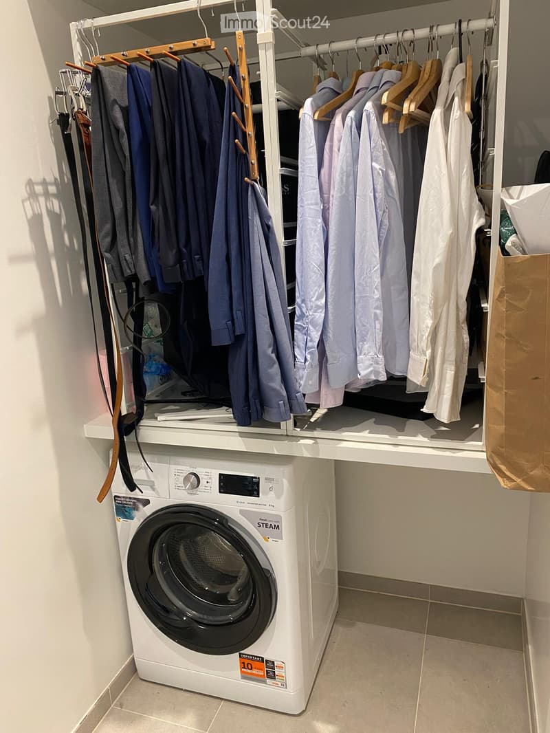 A modern bathroom with a white washing machine and a built-in clothes rack holding various shirts and jackets.