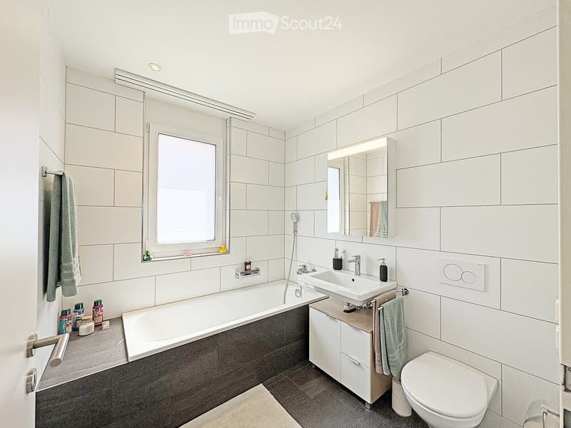 Modern bathroom with white tiles, bathtub, sink, toilet, mirror, and towel. Features a window with a small shelf holding toiletries and a decorative duck toy.