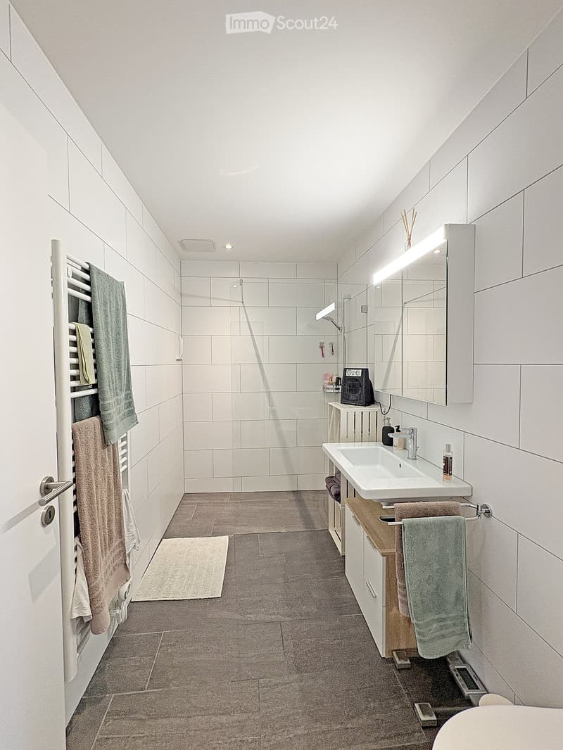 Modern bathroom with white tiles, a double sink, a large mirror with built-in lighting, a towel rack with towels, a shower area, and a small cabinet.