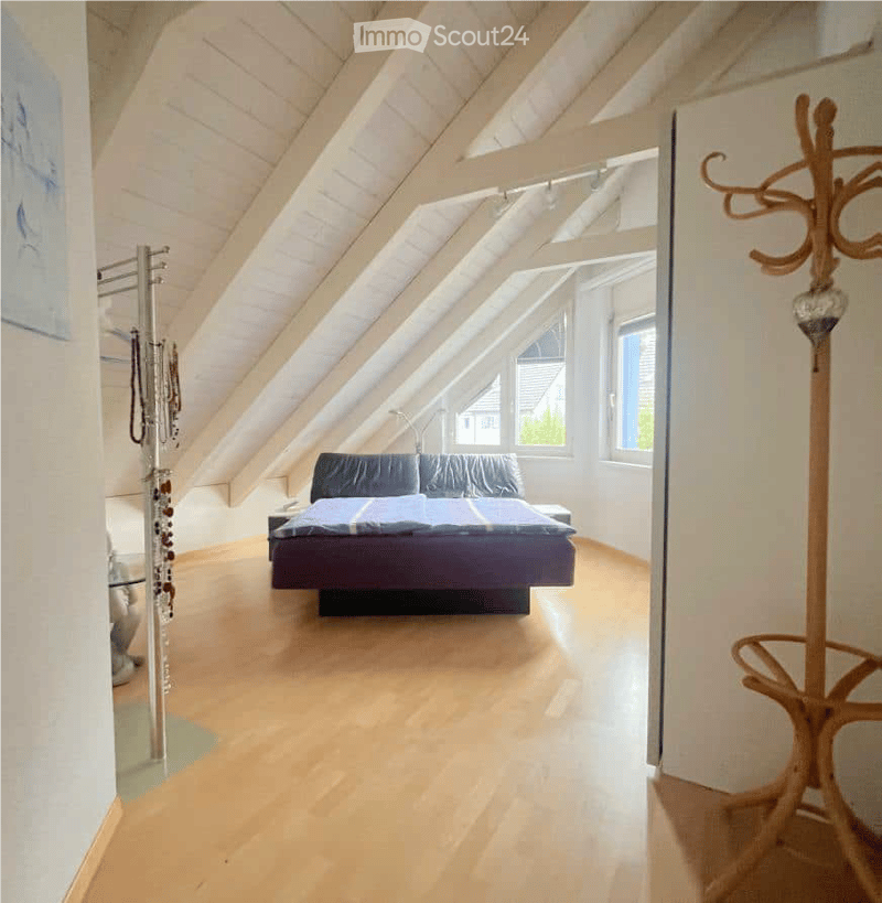 Bright attic bedroom with wooden floors, slanted white ceilings, large windows, and a bed with blue bedding.