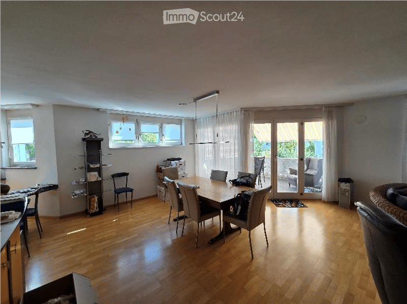 Bright, open-plan dining and living area with wooden flooring, large windows, and sliding glass doors leading to a balcony. Features a dining table with chairs, a sofa, a shelf, and a hanging lamp.