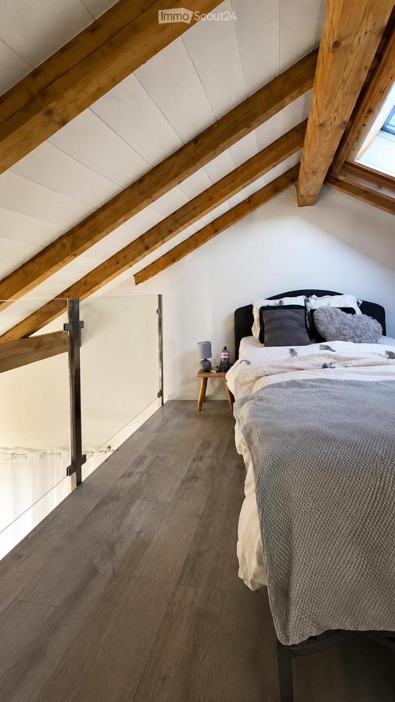 Cozy attic bedroom with wooden beams, slanted ceiling, and skylight. Features a bed with pillows and blankets, a nightstand with a lamp, and a small wooden stool.