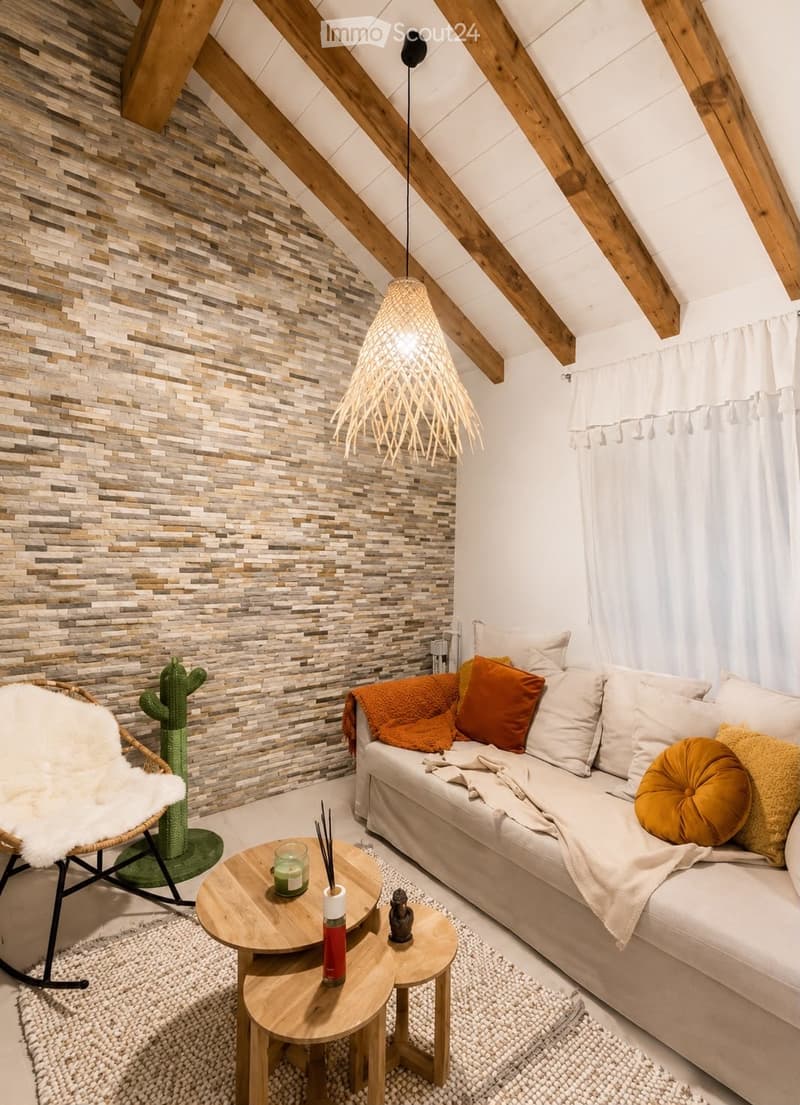 A cozy living room with exposed wooden beams, a textured stone accent wall, a white sofa with orange and yellow cushions, a wooden coffee table, a wicker chair with a white fur throw, and a hanging woven pendant light.