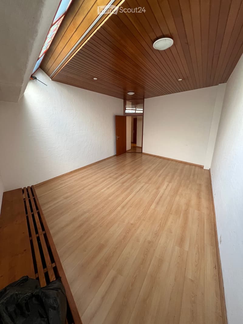 Empty room with wooden flooring, white walls, and a wooden ceiling with recessed lighting. A door leads to another room, and a window is visible at the top left corner.