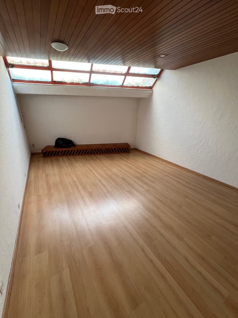 Empty room with wooden flooring, white walls, and a small bench with a bag on it. The ceiling has wooden paneling and a skylight window.