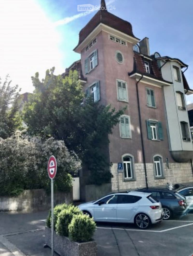 Multi-story residential building with pink facade, multiple windows, and a small garden area in front.