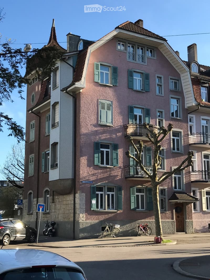 A multi-story residential building with pink facade, green shutters, and multiple windows. Features a balcony on the second floor and a tree in front.