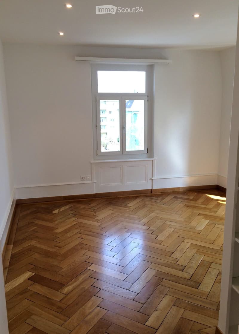 Empty room with herringbone-patterned wooden flooring, white walls, and a large window with double doors leading outside.