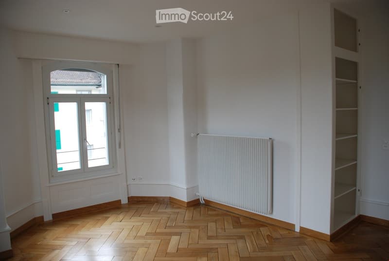 Empty room with wooden parquet flooring, white walls, a radiator, and a built-in shelf unit. The room has large windows allowing natural light to enter.