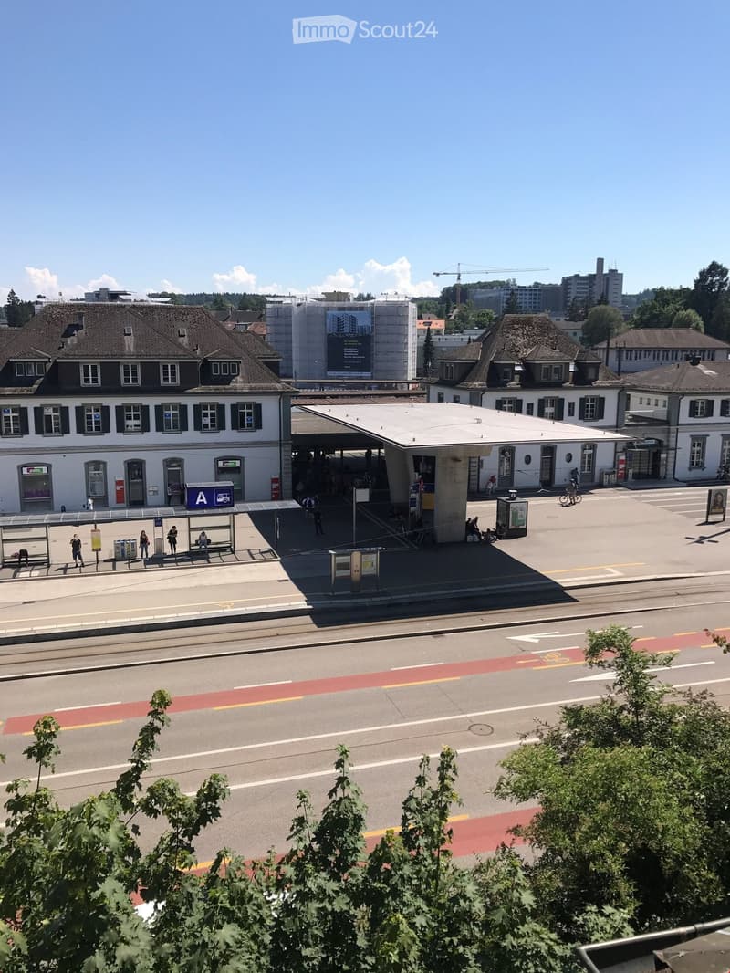 A train station with a modern platform shelter, a bus stop, and surrounding buildings with traditional architecture.