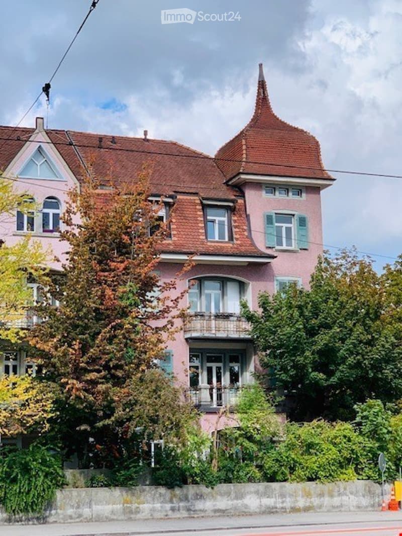A pink multi-story building with a red-tiled roof, decorative gables, and multiple windows, surrounded by trees and greenery.
