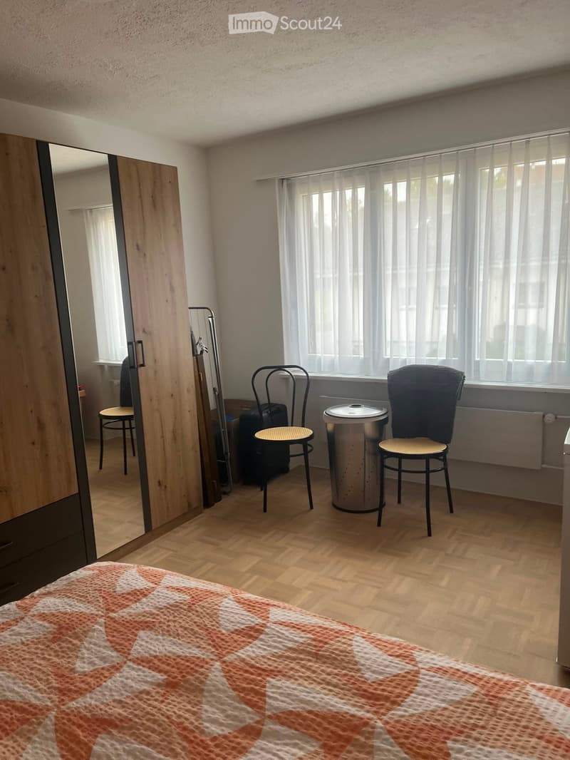 Bedroom with wooden flooring, a bed with an orange and white patterned bedspread, a small round table with two chairs, a wardrobe with a mirror, and a suitcase near the window.