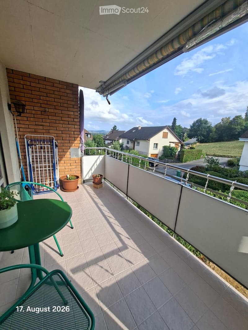 Balcony with tiled floor, green table and chairs, potted plants, and a view of the surrounding area.