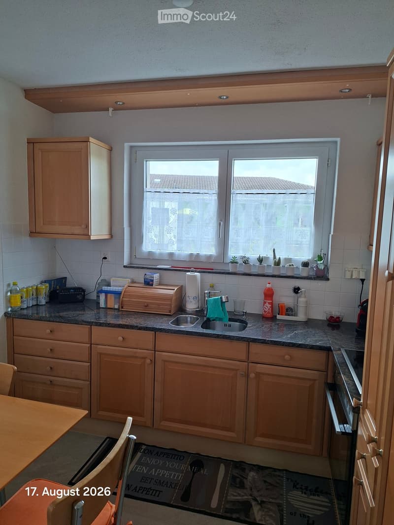 A well-lit kitchen with wooden cabinets, granite countertops, a sink, and various appliances and items on the counter, including a radio, bottles, and a dish rack.