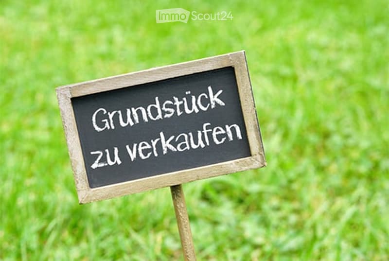 A small wooden signboard with the text "Grundstück zu verkaufen" (Land for sale) placed on a wooden stake in a grassy field.