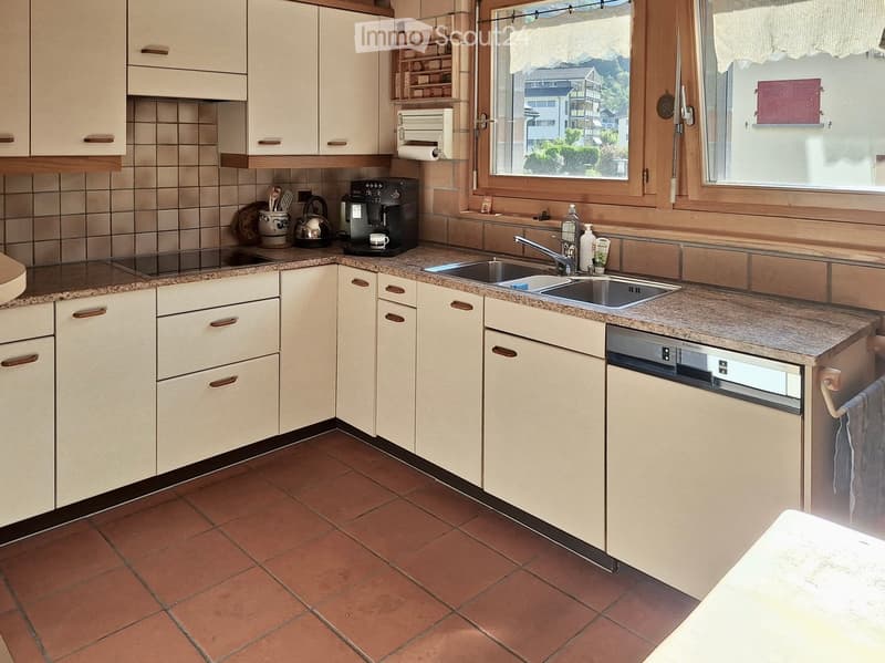 Bright kitchen with cream-colored cabinets, granite countertops, and a double sink. Appliances include a coffee maker, teapot, and dishwasher. The room has tiled floors and large windows offering a view of the surroundings.