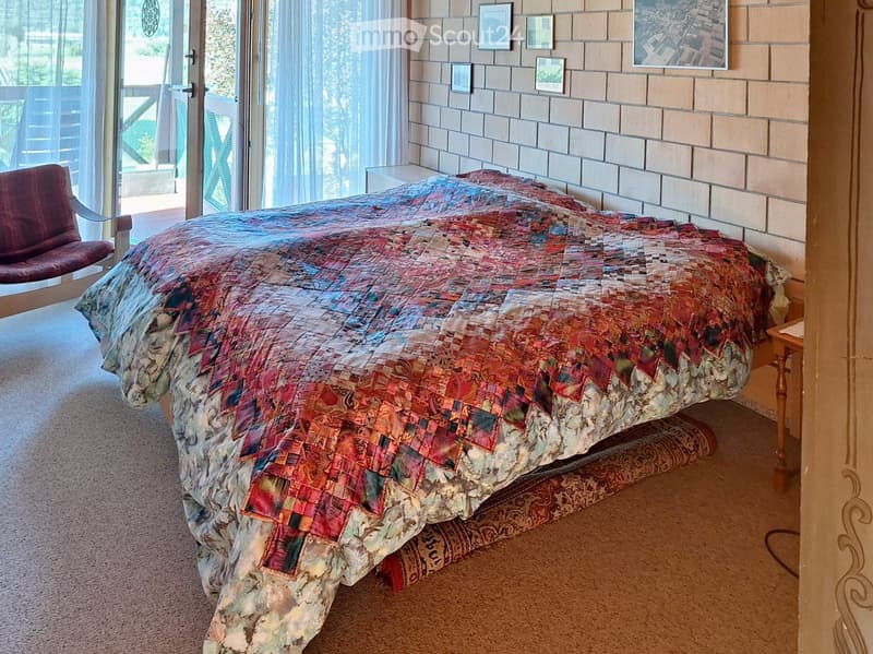 Bedroom with a large bed covered in a colorful patchwork quilt, a wooden nightstand, and a red chair near a large window with sheer curtains.