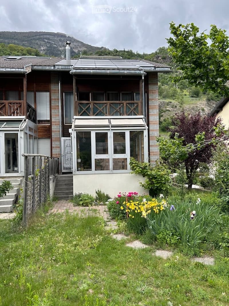 Two-story house with a balcony, surrounded by a garden with colorful flowers and a mountain view in the background.